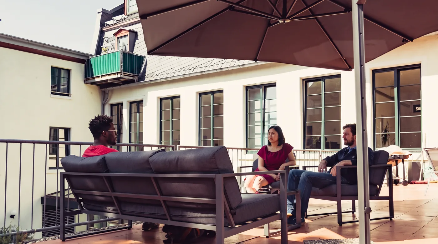 Drei Personen sitzen auf einer sonnigen Dachterrasse unter einem Sonnenschirm und unterhalten sich.