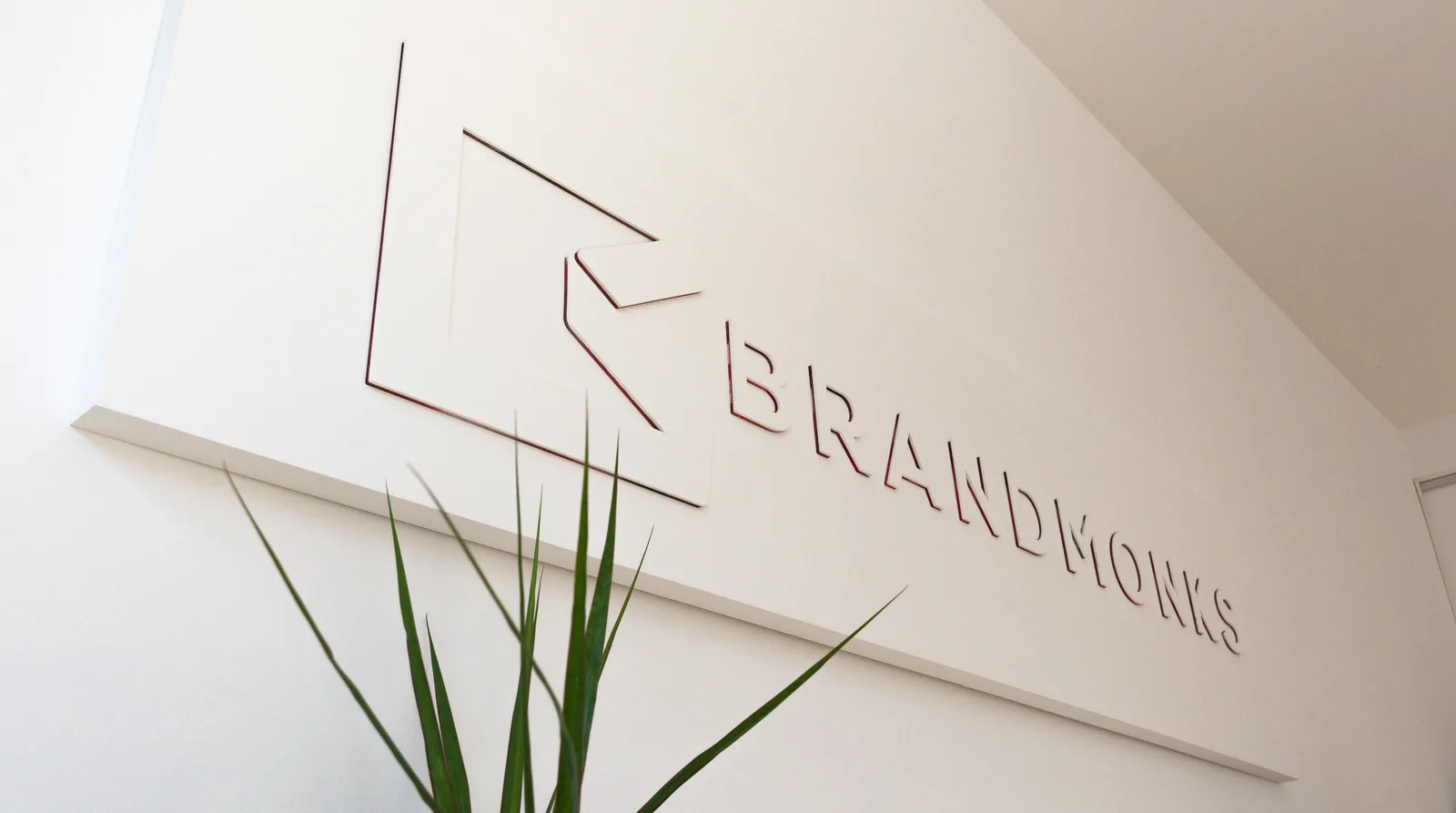Brandmonks-Schriftzug und Logo als Relief an einer hellen Wand, davor eine Pflanze.