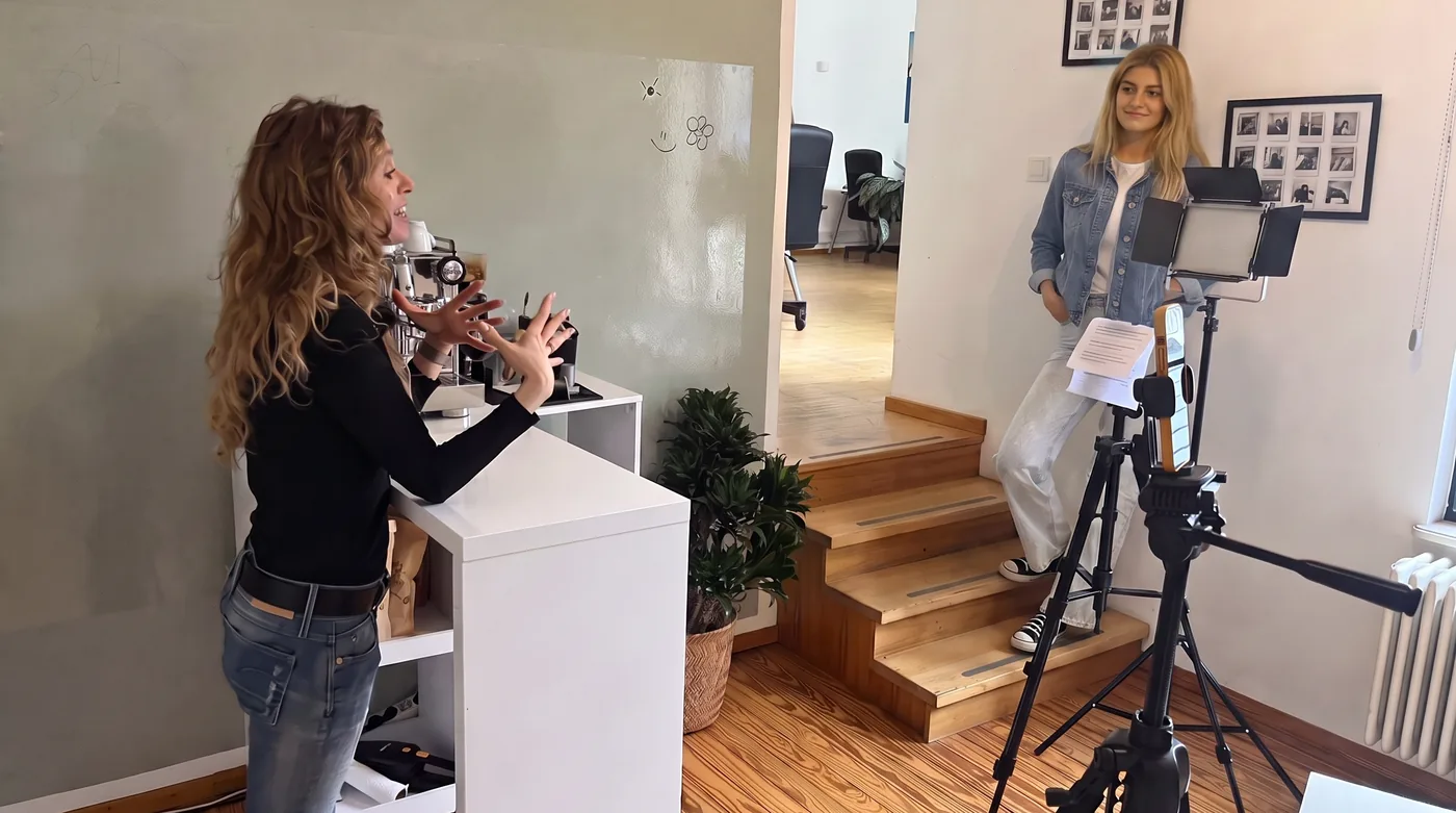 Zwei Personen in einem Büro bei einer Videoaufnahme: eine spricht vor der Kamera auf dem Stativ, die andere steht an der Theke.