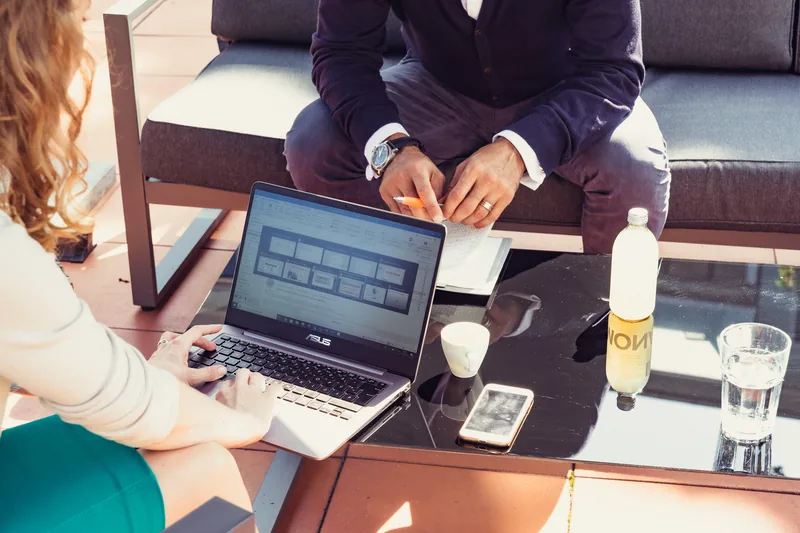 Zwei Personen arbeiten im Freien an einem Tisch, eine am Laptop, die andere macht Notizen.