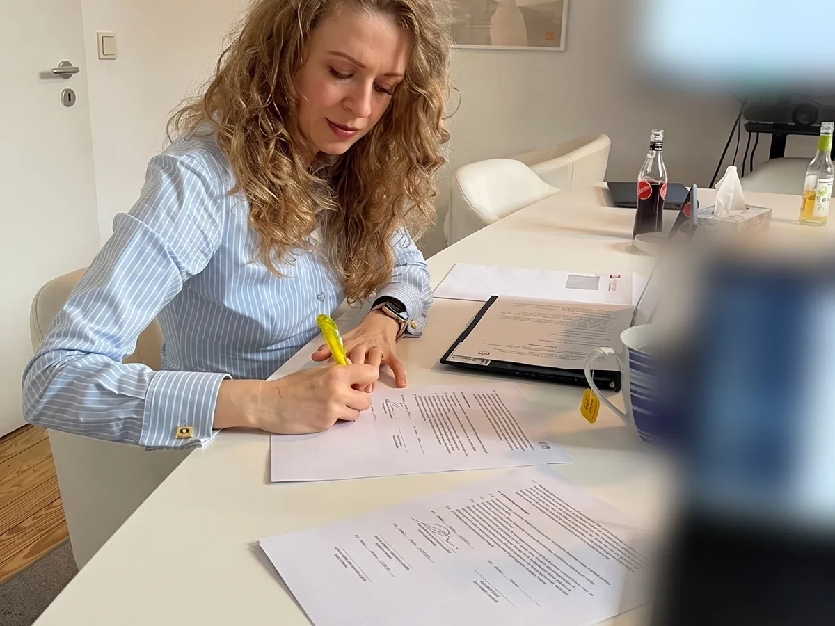Frau an einem Besprechungstisch markiert eine Stelle in einem ausgedruckten Dokument; daneben liegen weitere Unterlagen und eine Mappe.