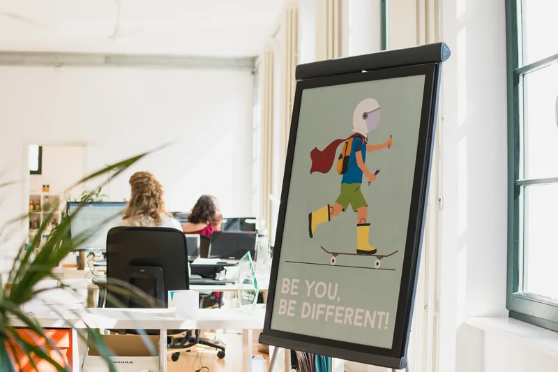 Blick in ein helles Büro mit Arbeitsplätzen, im Vordergrund ein Plakatständer mit dem Leitsatz „Be you, be different“.