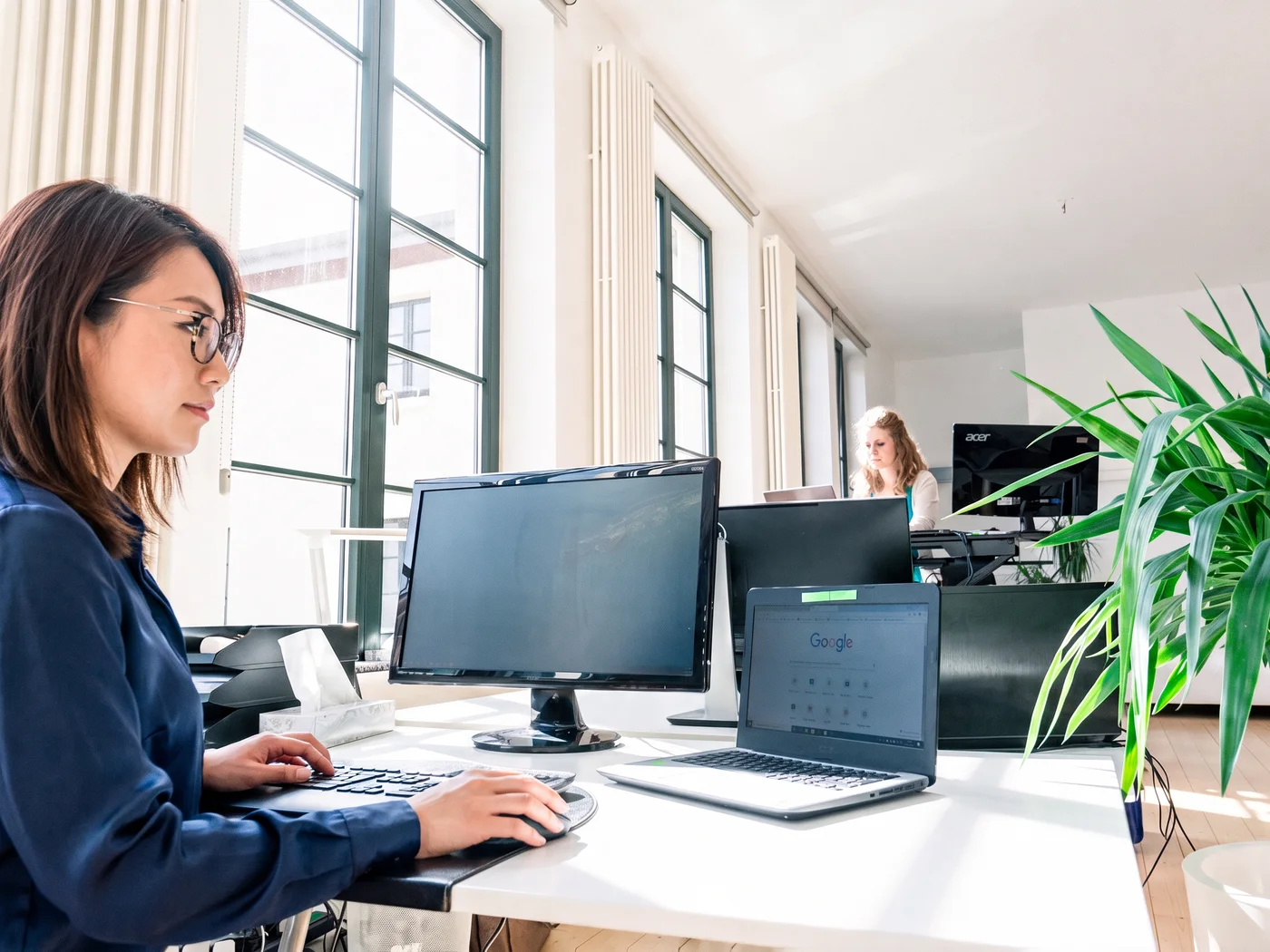Frau mit Brille arbeitet an einem Schreibtisch mit Bildschirm und Laptop in einem hellen Büro, im Hintergrund ein zweiter Arbeitsplatz.