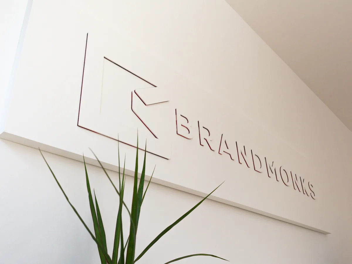 Brandmonks-Schriftzug und Logo als Relief an einer hellen Wand, davor eine Pflanze.